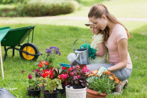 A woman and a young girl are in a well-maintained garden with lush, green grass, surrounded by blooming flower pots containing vibrant red, pink, purple, and orange flowers. They are kneeling together on the lawn, engaging in planting or tending to the flowers, with gardening gloves on. Behind them, there is a green wheelbarrow with yellow wheels and a garden hose reel, indicating outdoor garden care activities. The garden features a neatly trimmed hedge in the background, and the scene is illuminated by natural daylight, suggesting a bright, clear day. The setting reflects a typical back garden in Greenwich, ideal for outdoor gardening and lawn maintenance, consistent with professional gardening services offered in the area by Gardening Services Greenwich.