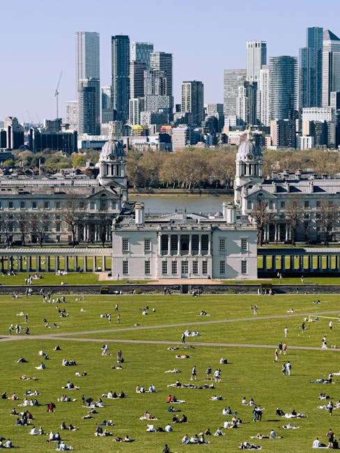 A large urban park with expansive, neatly maintained green lawns where numerous people are sitting, lying, and relaxing on the grass, enjoying clear weather. The park features wide pathways crisscross