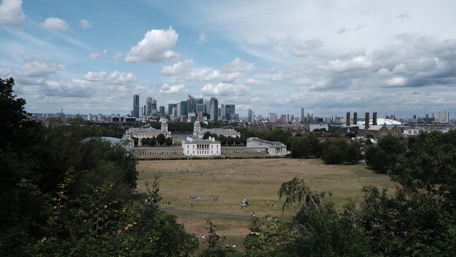 A panoramic view of a large, open park with expansive grassy areas and scattered trees, set against a backdrop of a modern city skyline featuring tall skyscrapers and industrial buildings under a part