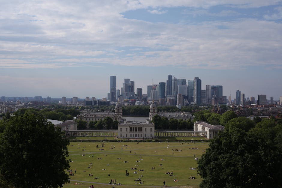 A panoramic view of a large, open park with expansive grassy areas and scattered trees, set against a backdrop of a modern city skyline featuring tall skyscrapers and industrial buildings under a part